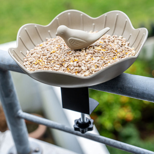Olijfgroene voederschaal met klem aan een balkonreling, gevuld met vogelzaad